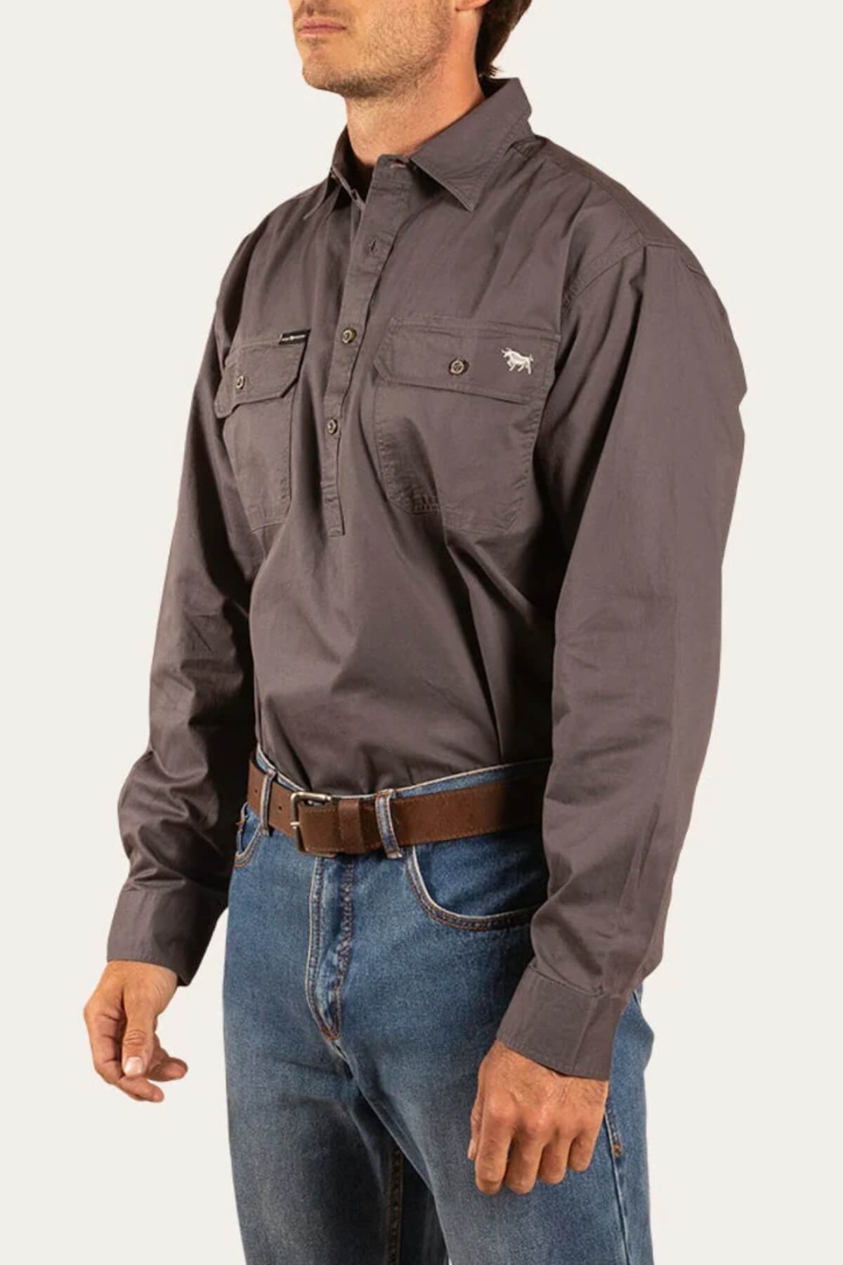 Someone wearing a Ringers Western King River Half Button Shirt in dark gray with long sleeves and two chest pockets, paired with blue jeans and a brown belt, stands against a plain light background.
