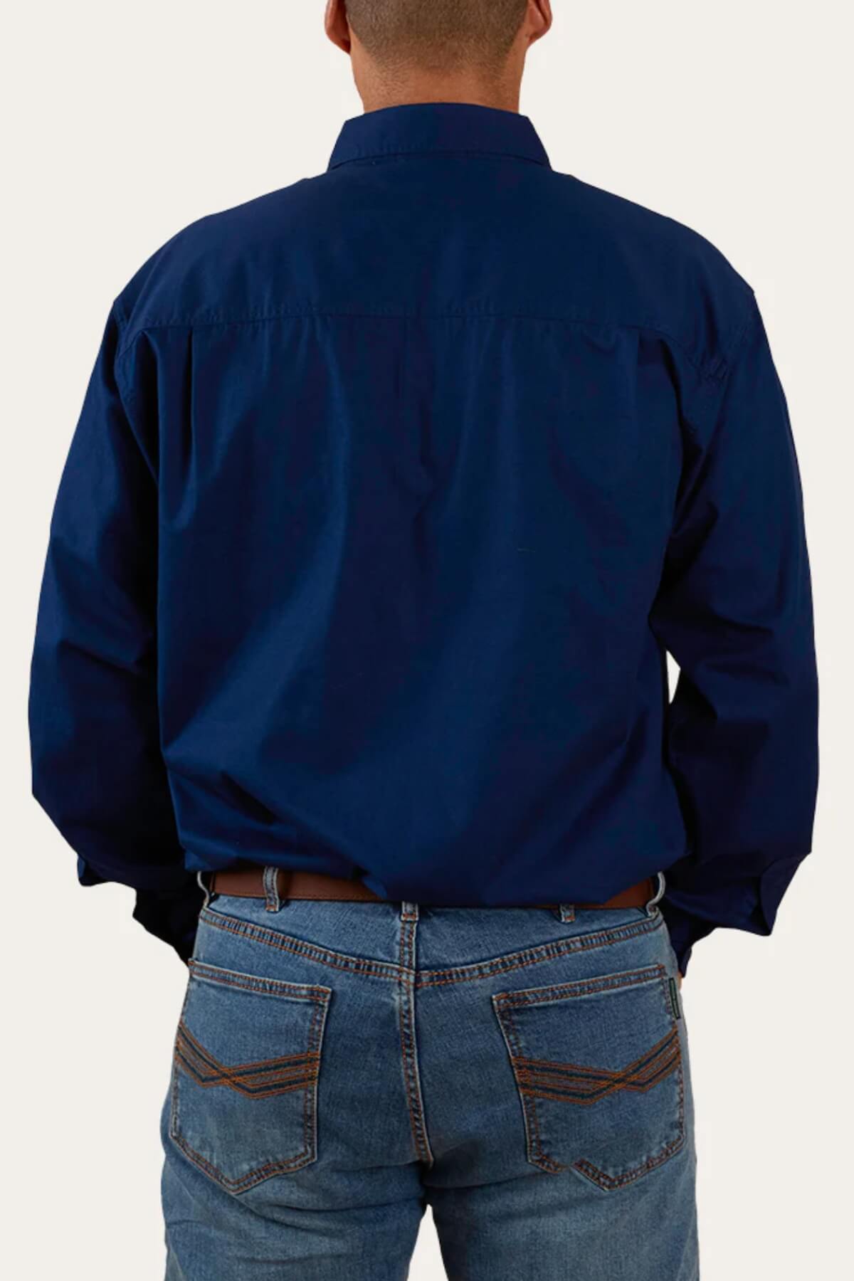 A person wearing a dark blue Ringers Western King River Half Button 100% cotton long-sleeve work shirt and blue jeans is shown from the back. The shirt is untucked, and the jeans have visible stitching on the back pockets. The person's hands are in their pockets, against a plain, light background.