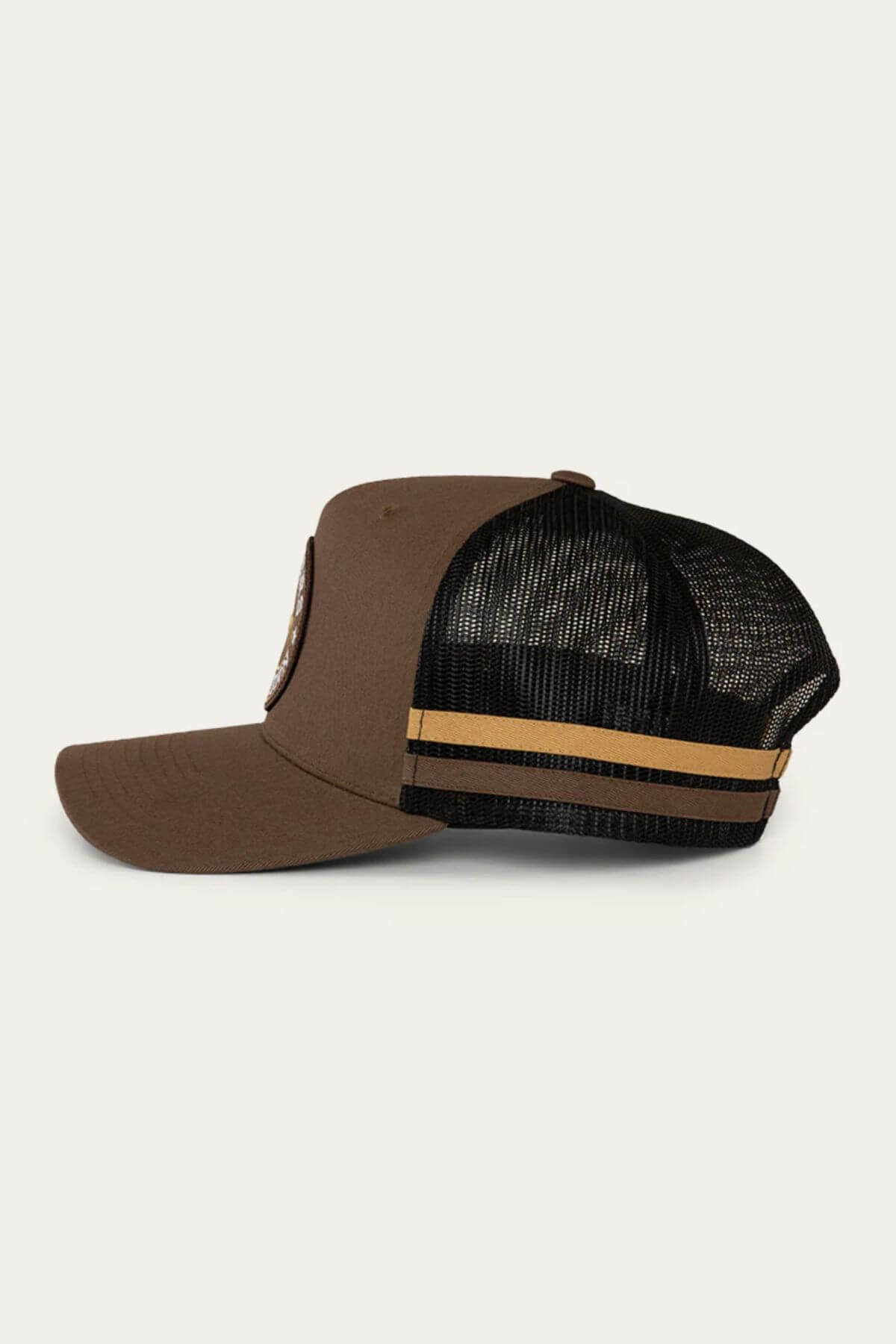 Side view of the Ringers Western McCoy Trucker Cap in brown cotton twill, featuring a black mesh back with two yellow stripes. It has a circular embroidered patch on the front panel and a classic curved bill.