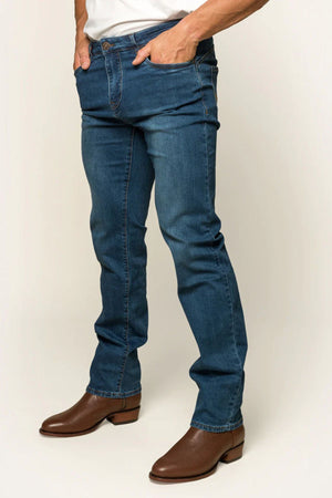 Wearing Ringers Western Mitchell Men's Straight Leg Jeans, a white shirt, and brown leather boots, a person stands with hands in pockets against a plain background, epitomizing the classic midrise straight-leg denim style of Ringers Western fashion.