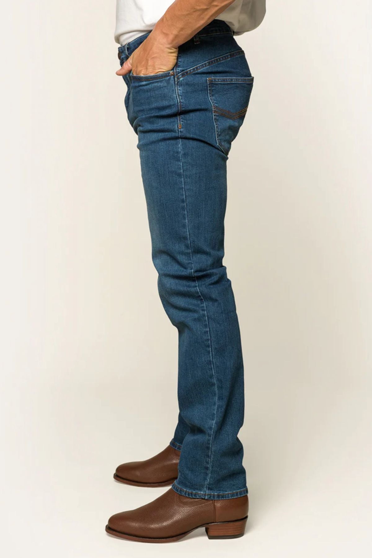 A person stands sideways wearing Ringers Western Mitchell Mens Straight Leg Jeans and brown boots, with one hand in the front pocket. The background is a plain light color, exuding timeless style.