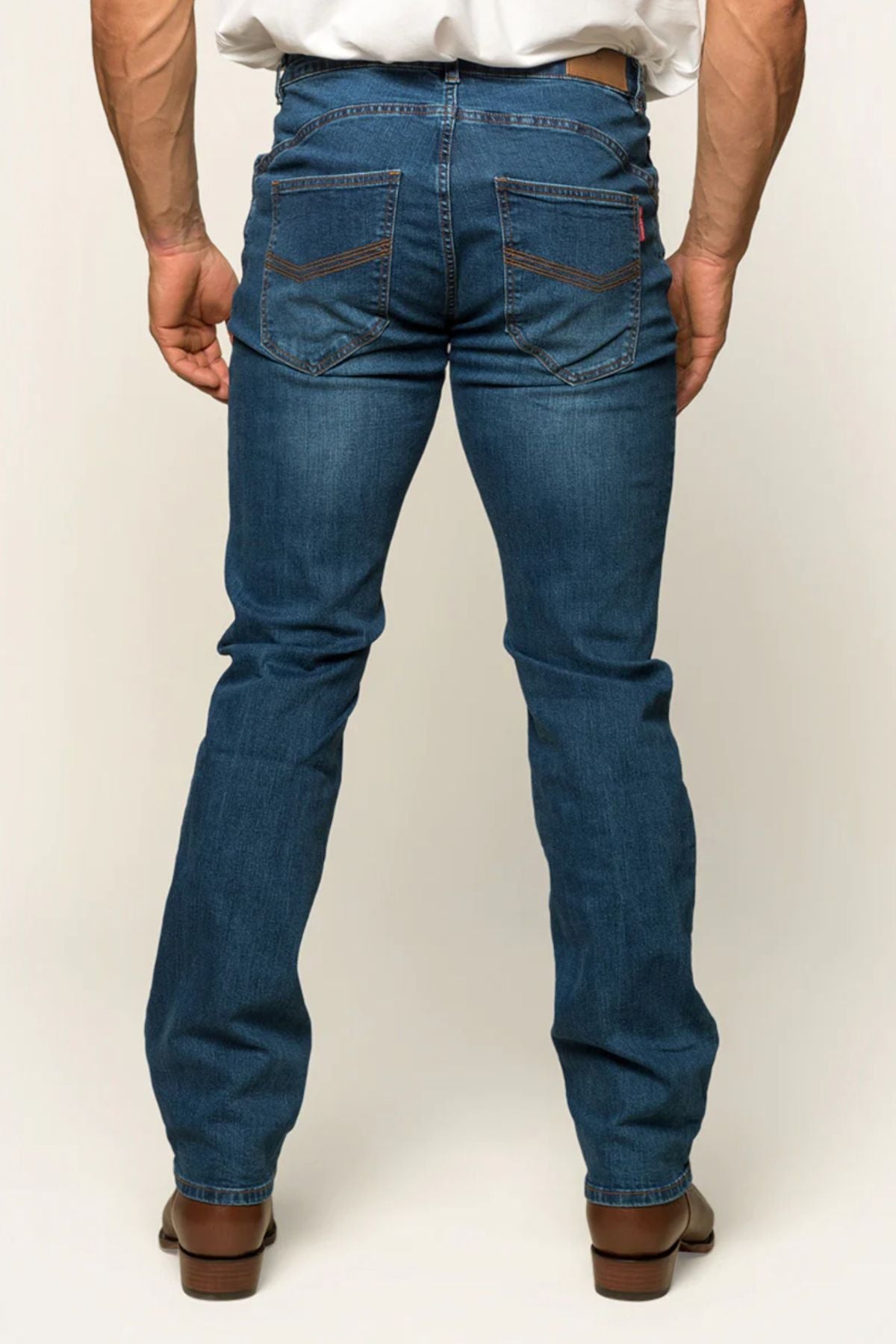 A person in Ringers Western Mitchell Mens Straight Leg Jean, midrise straight leg design, with a white shirt accenting back pockets and brown stitching. Sporting brown shoes against a neutral backdrop, they embody Ringers Western's laid-back style.