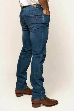 A person wearing Ringers Western Mitchell Mens Straight Leg Jean in blue denim and brown leather boots is standing with their right side visible. The jeans have distinctive stitching on the back pockets, and their midrise straight leg cut pairs perfectly with a tucked-in white shirt.