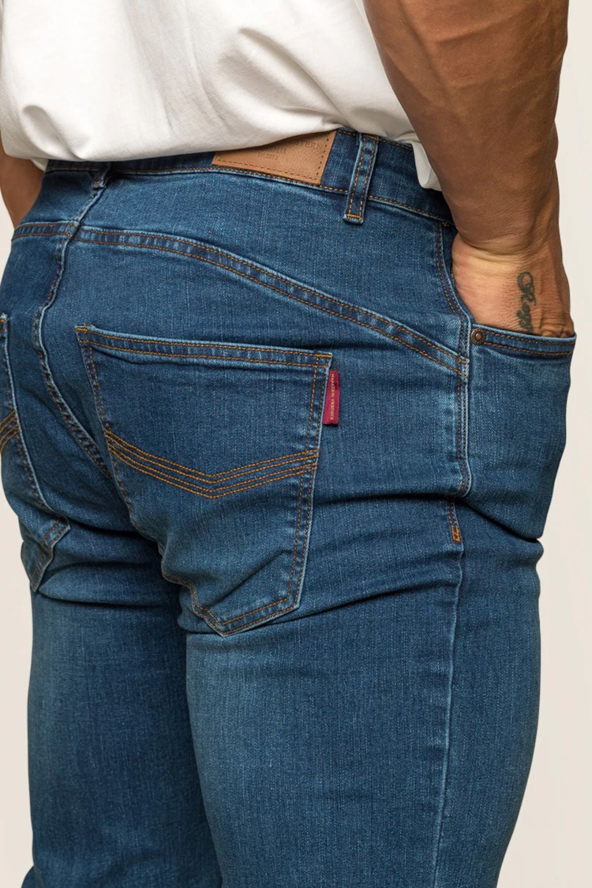 A close-up showcases a person in Ringers Western Mitchell Men's Straight Leg Jeans, featuring distinctive stitching and a small red tag on the back pocket. They wear a white shirt, with their right hand in the pocket, revealing an arm tattoo that complements the midrise straight leg style.
