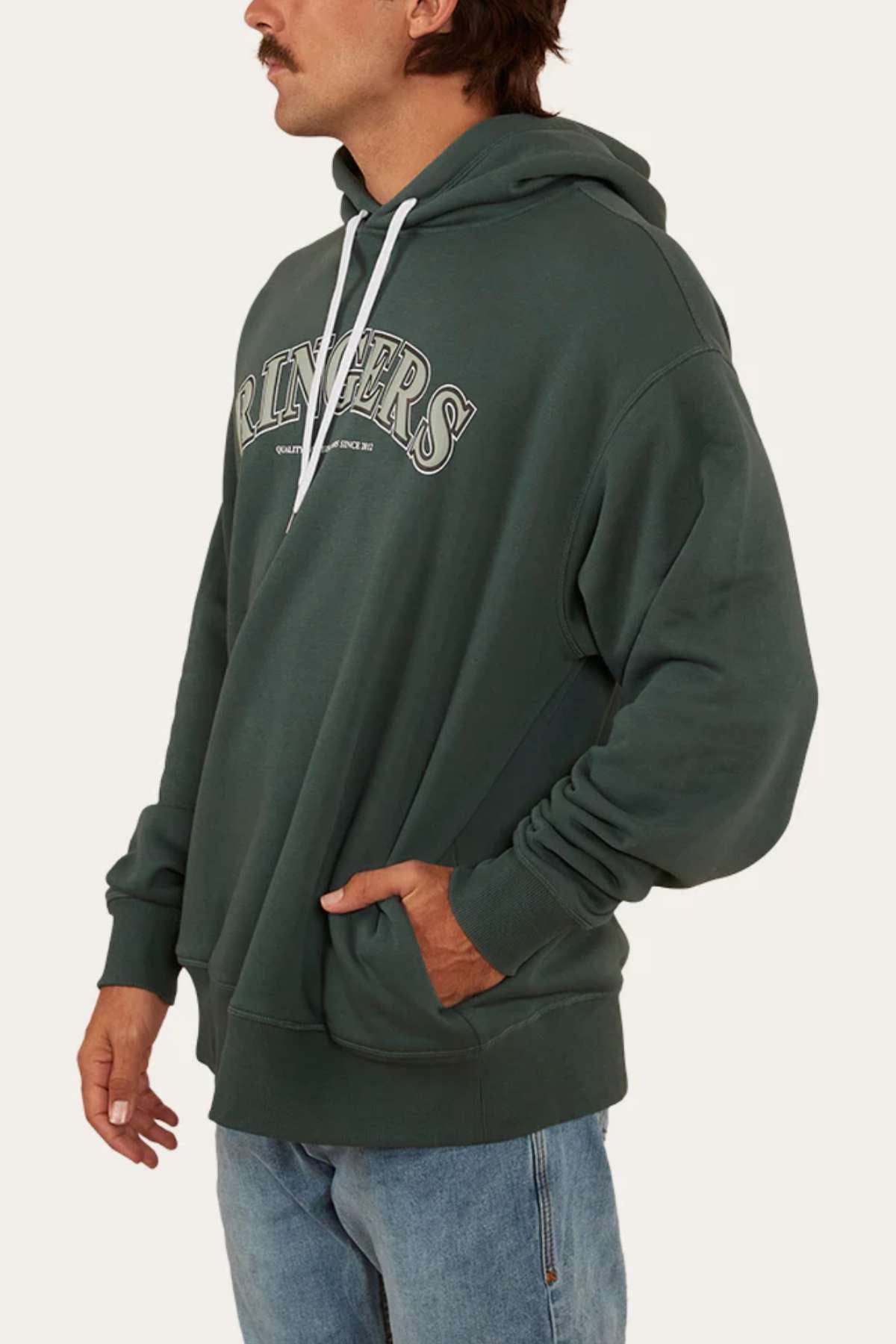 A person with a mustache is wearing a dark green, oversized fit Ringers Western Princeton Hoodie in brushed fleece with "Ringers" printed on the front, paired with light blue jeans. The hoodie features white drawstrings and the individual has one hand in the front pocket.