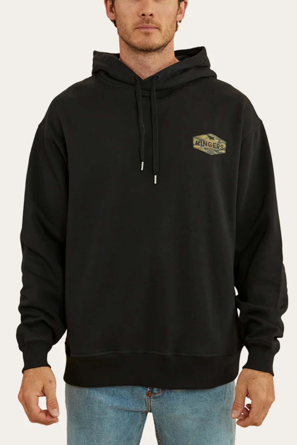 A man wearing a Ringers Western Servo Hoodie in black, featuring a small "Ringers" logo on the left chest, stands facing forward. The brushed fleece fabric and oversized fit offer both comfort and style, complementing his light blue jeans perfectly.