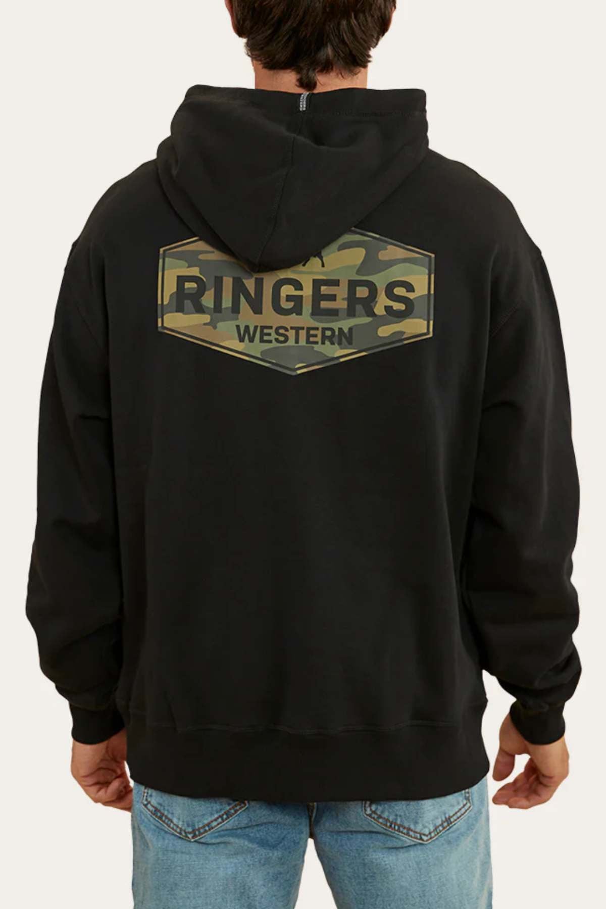 A person wearing a Ringers Western Servo Hoodie, which features a black base with a green camouflage design and the text "Ringers Western" on the back. The individual is dressed in blue jeans, facing away from the camera with the hood down. The oversized fit and brushed fleece material enhance their relaxed appearance.