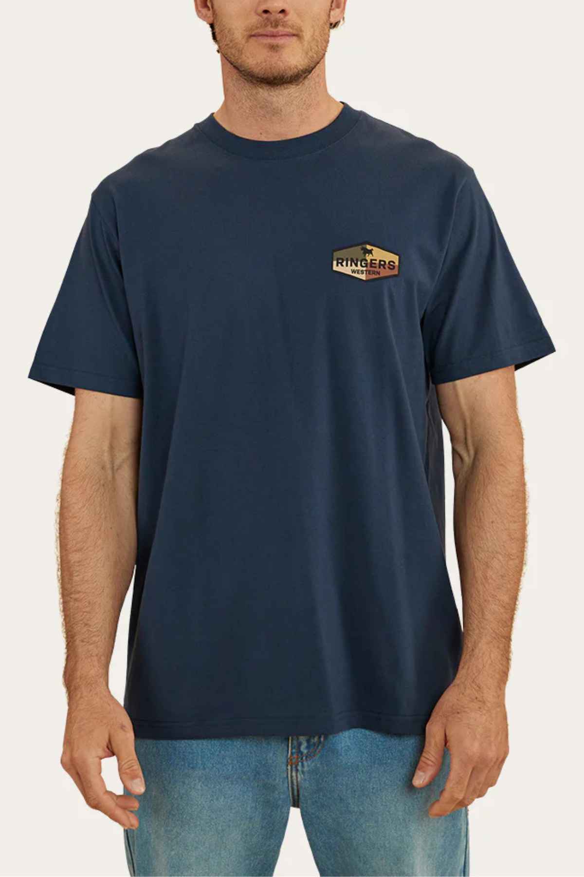 A person is wearing a Ringers Western Servo Loose Fit T-Shirt in navy blue, crafted from 100% cotton jersey, paired with blue jeans. The shirt features the brand's logo on the left chest against a plain off-white background.
