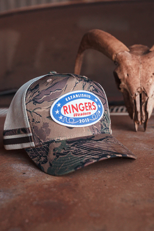 The Ringers Western Shooter Deep Fit Trucker Cap, featuring camouflage mesh, a blue, red, and white "Ringers Western" patch, and adjustable snapback closure, sits on a rusty surface with a horned skull blurred in the background.