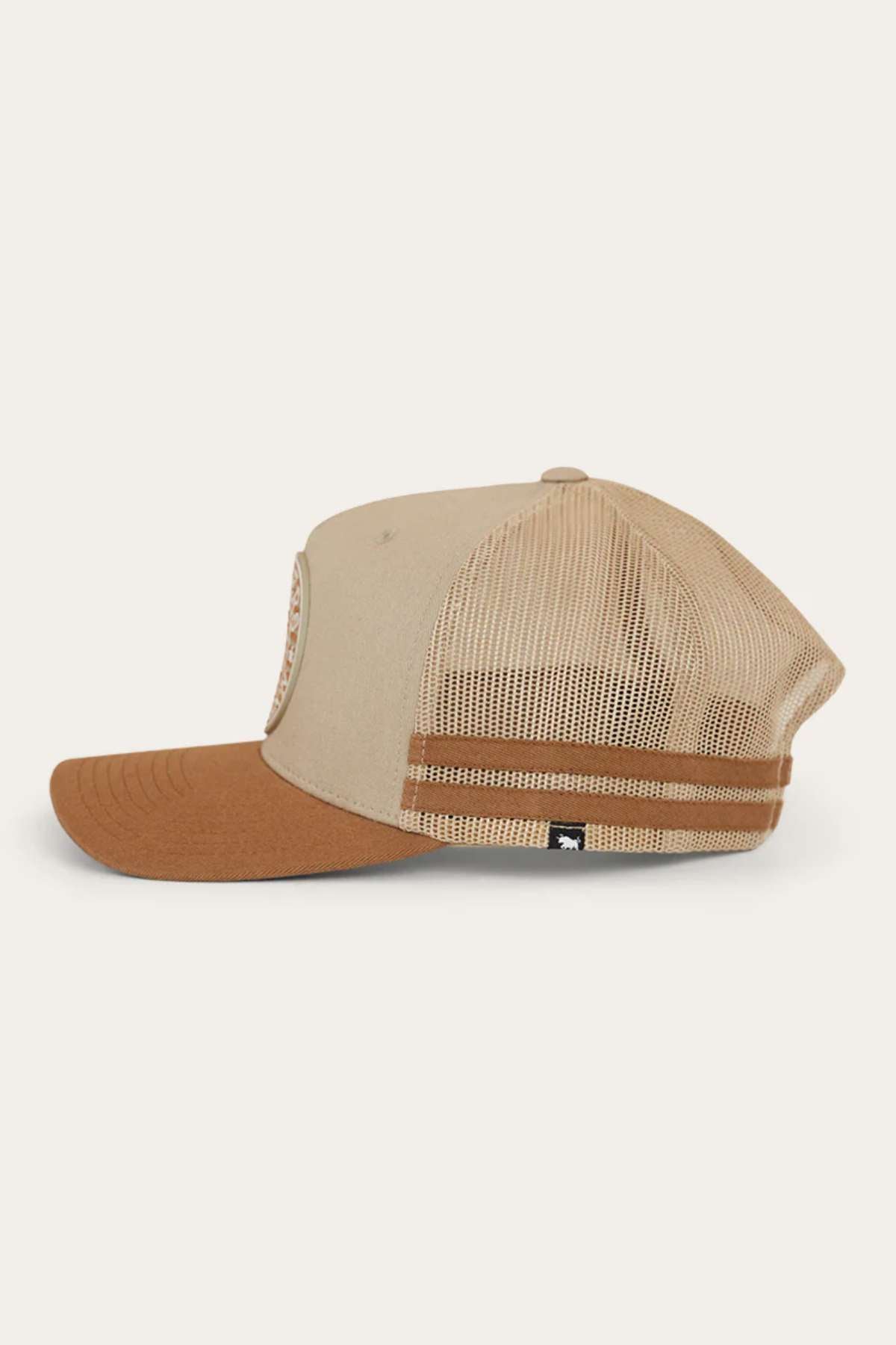 A side view of the Ringers Western Signature Bull Flag Trucker Cap, featuring a beige and brown color scheme with a curved brown brim. The back includes a beige mesh with an adjustable snapback closure and a small black tag on the lower portion. The front showcases partially visible stitching details.