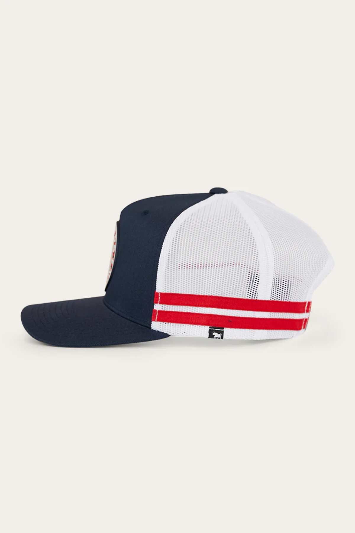 The Ringers Western Signature Bull Flag Trucker Cap features navy blue and white mesh, two red stripes, a small black tag above the adjustable snapback closure, and is shown from the side on a plain light background.