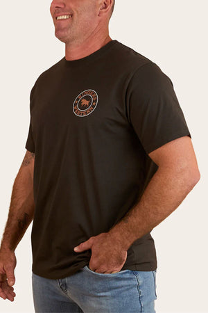 A smiling man wears the Ringers Western Signature Bull Loose Fit T-Shirt, 100% cotton with a small circular logo on the left chest, paired with light blue jeans, standing against a plain light background.