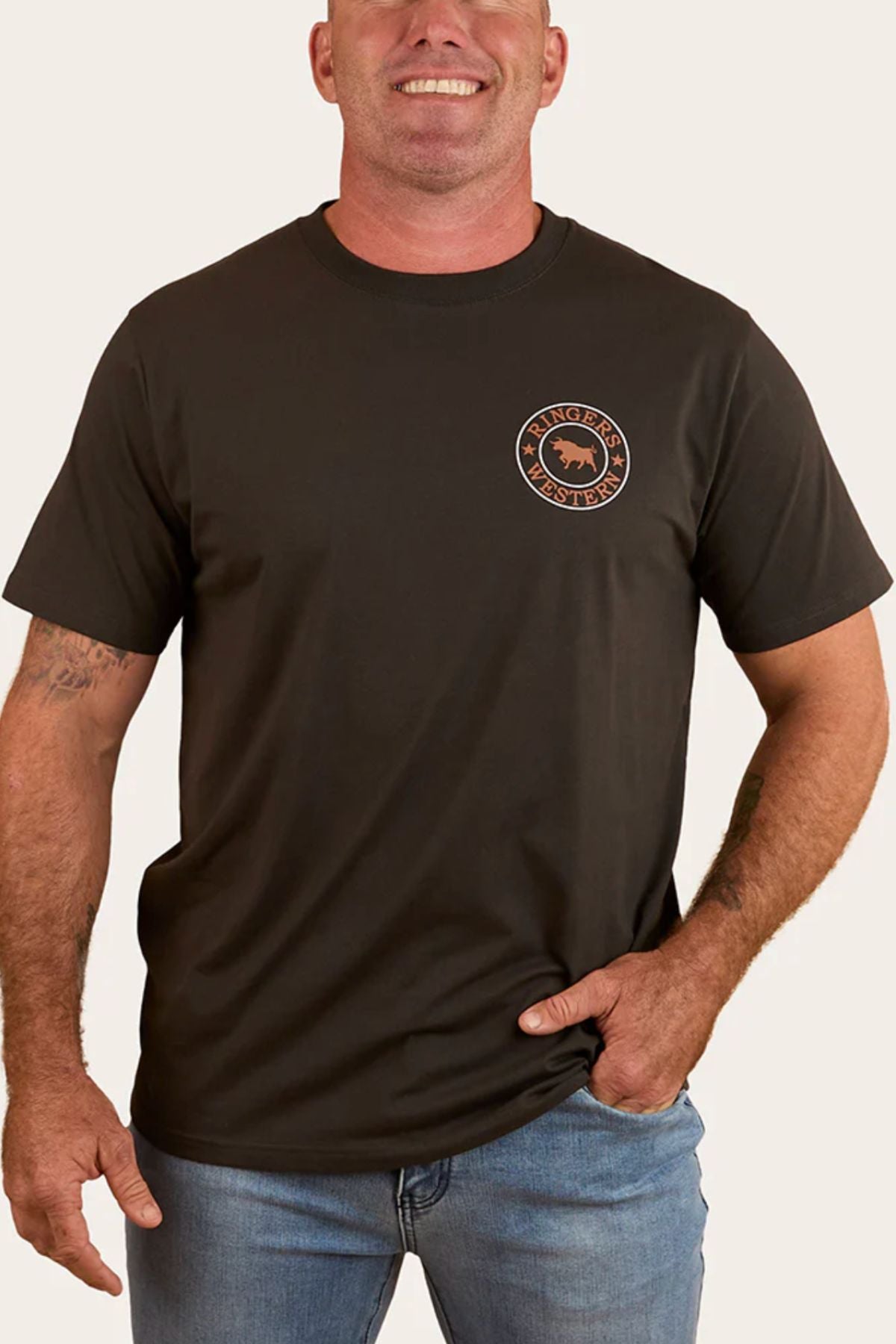 A smiling man wears a dark Ringers Western Signature Bull Loose Fit T-Shirt with a round orange and black bull logo, paired with light blue jeans, standing casually with one hand in his pocket.