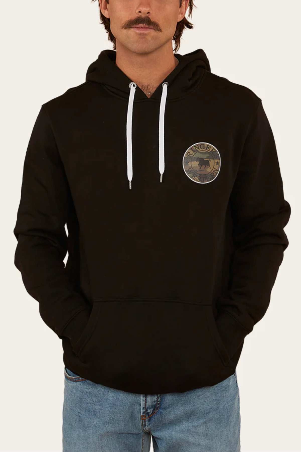 A person with a mustache, wearing the Ringers Western Signature Bull Pullover Hoodie in relaxed-fit black featuring a round emblem on the chest and white drawstrings, paired with blue jeans from Ringers Western. They stand against a plain white background with hands in the hoodie pockets.