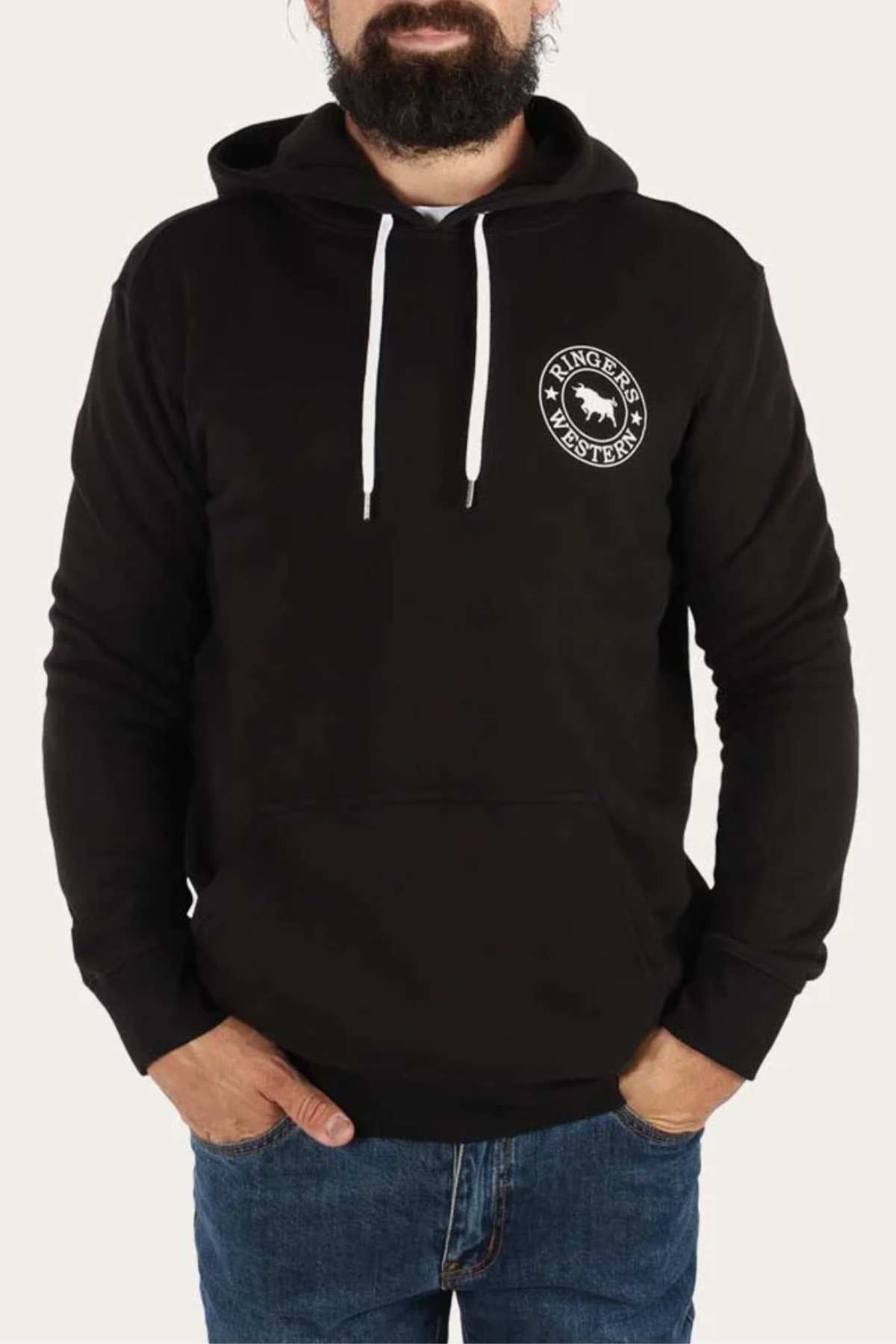 A bearded man is wearing the Ringers Western Signature Bull Pullover Hoodie, featuring a black casual design with a white drawstring and a round logo on the left chest that reads "Ringers Western" with a cow graphic in the center. He has his hands in the front pocket of the relaxed fit hoodie and is also wearing blue jeans.