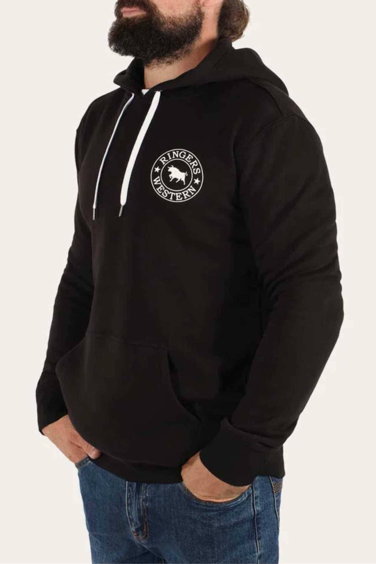 A bearded man is seen wearing a black Ringers Western Signature Bull Pullover Hoodie, characterized by its relaxed fit and white drawstrings. The hoodie features a prominent "Ringers Western" circular logo on the left chest area. He has his left hand casually tucked into the front pocket of the hoodie and is paired with blue jeans. The background is plain white.
