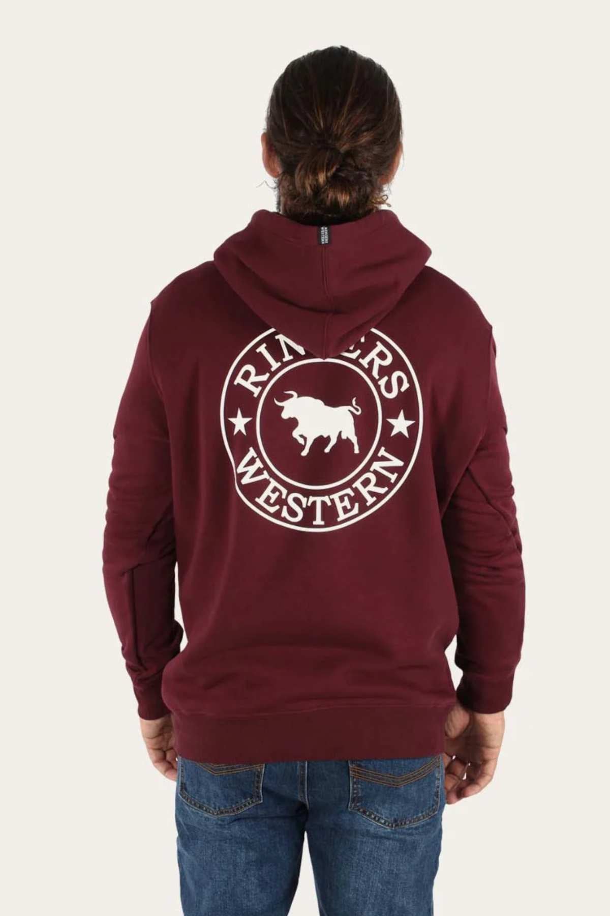 A person with long hair tied back is seen from behind, wearing a maroon Ringers Western Signature Bull Pullover Hoodie adorned with a large white logo and an image of a bull on the back. They are also dressed in blue jeans.