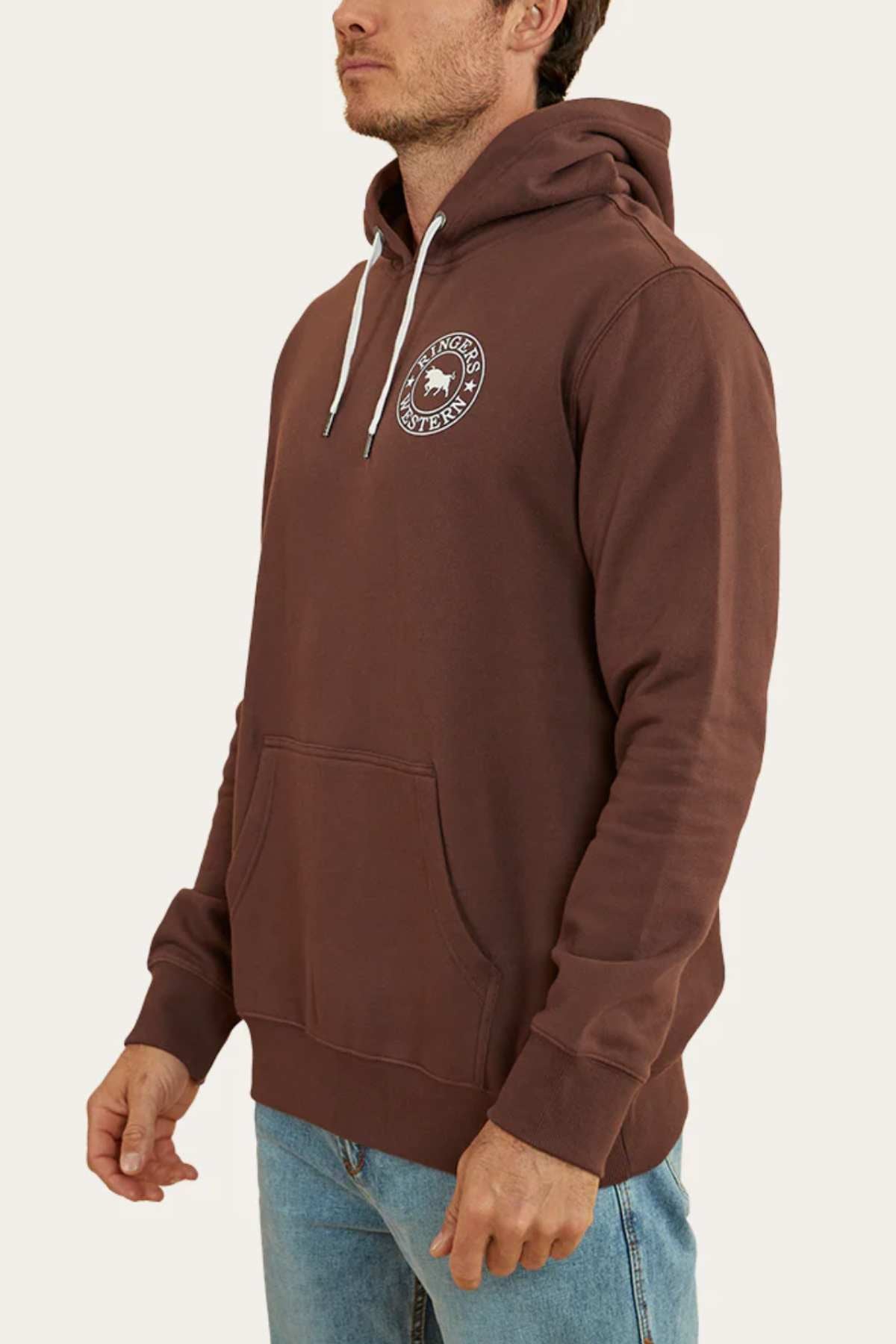 A person is wearing a comfortable Ringers Western Signature Bull Pullover Hoodie in brown, featuring a circular white logo on the left chest. The hoodie has white drawstrings and a front pouch pocket. The individual is also wearing light blue jeans and is standing sideways against a plain, light-colored background.