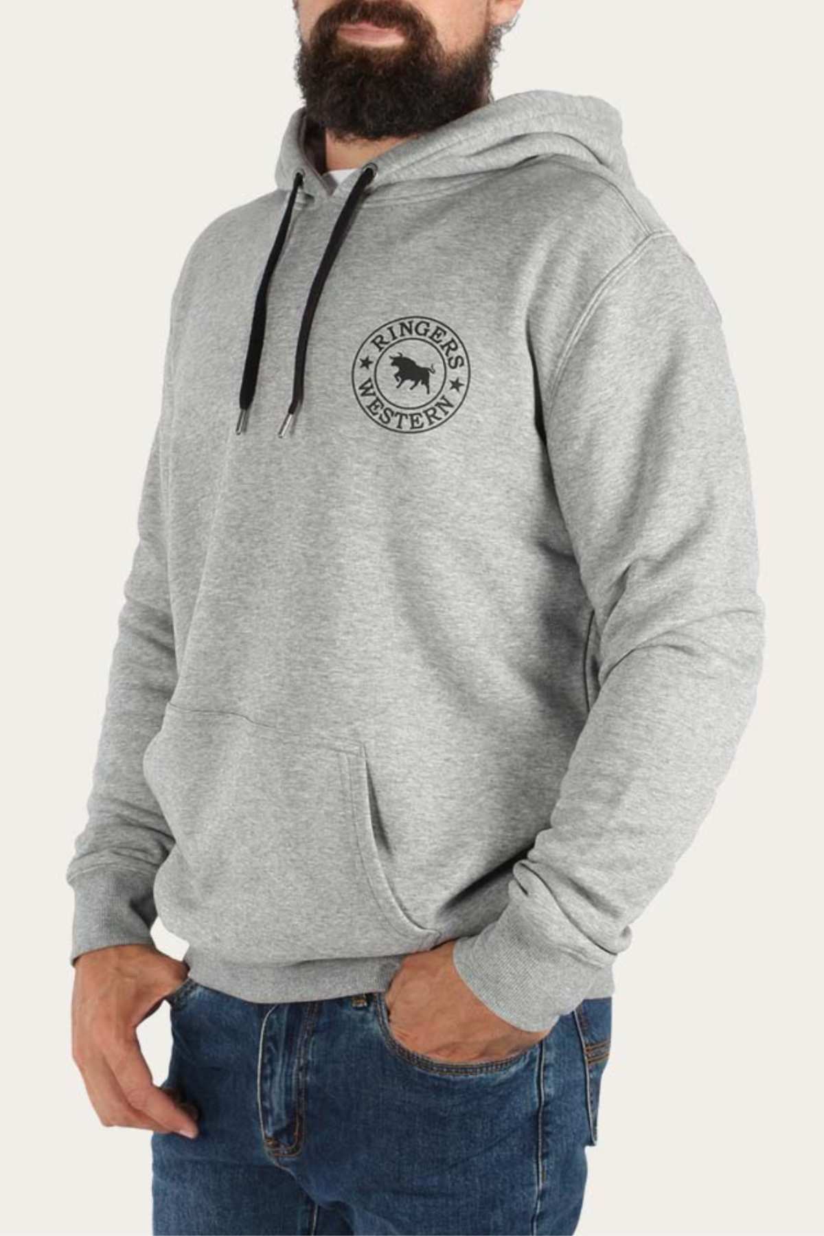 A bearded man is wearing a light gray Ringers Western Signature Bull Pullover Hoodie with a kangaroo pocket and dark drawstrings, paired with blue jeans. The comfortable hoodie features a black circular logo with a horse and stars on the left chest. He is standing against a plain white background.