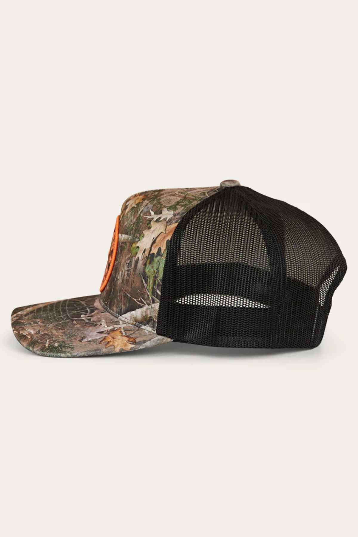 Side view of a Ringers Western Signature Bull Trucker Cap showcasing a black mesh back and orange logo. The cap's woodland pattern of leaves and twigs is set against a light cotton twill background.