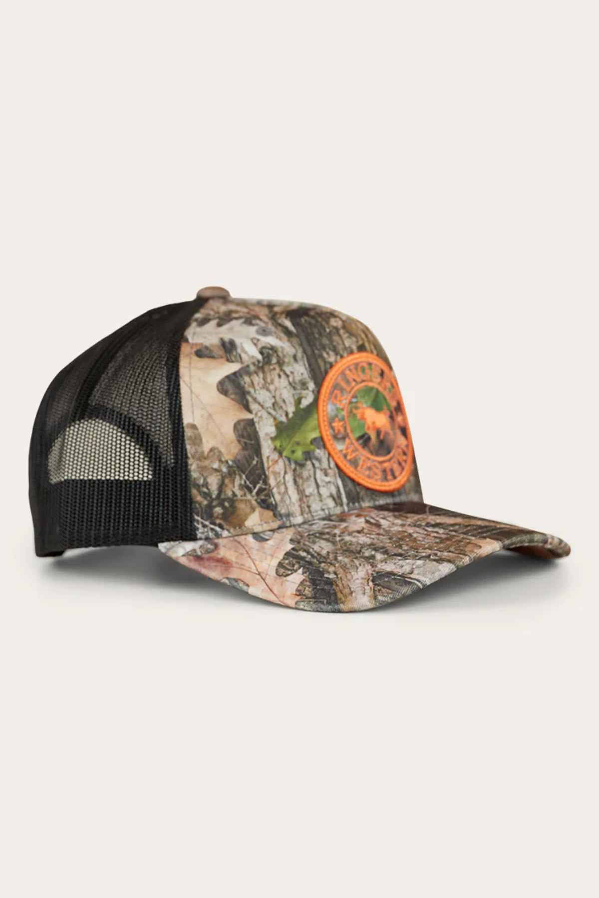 The Ringers Western Signature Bull Trucker Cap features a camouflage-patterned front and a black mesh back, adorned with an orange circular patch displaying a logo with text. Made from sturdy cotton twill, this cap is photographed on a light, neutral background, blending rugged style with comfort.