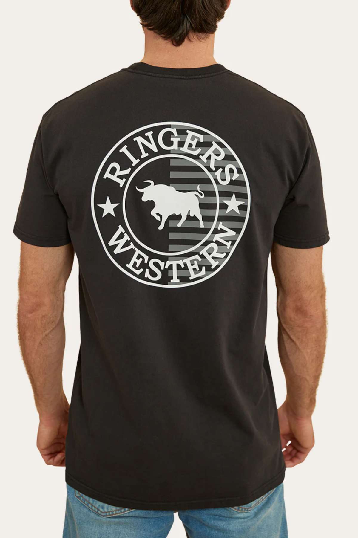A man, clad in blue jeans and facing away from the camera, is showcasing a black Ringers Western Signature Flag Stock Fit T-Shirt made from 100% cotton jersey. The shirt features a large circular logo on the back that reads "Ringers Western" with a bull silhouette in the center, surrounded by stars and stripes.