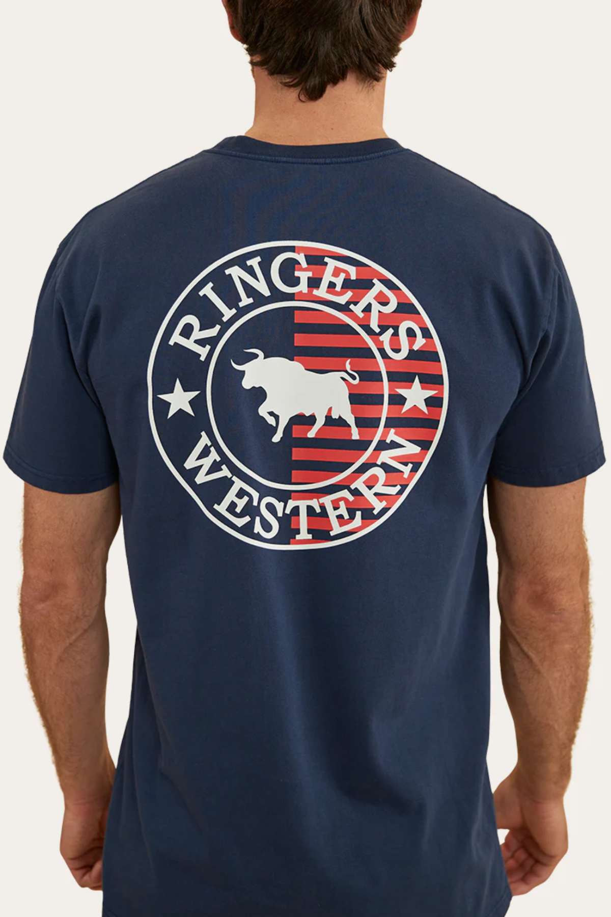 A person with short brown hair is seen from behind, wearing a dark blue Ringers Western Signature Flag Stock Fit T-Shirt made from 100% cotton. The t-shirt features a circular logo with a bull in the center and 'Ringers Western' written on it, accompanied by red and white stripes in the background.