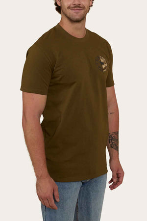 A person wearing the Ringers Western Signature Flag Stock Fit T-Shirt, a brown cotton jersey with a small circular logo on the chest, pairs it with light blue jeans. Standing against a plain background, their right arm slightly bent reveals a tattoo on the forearm.