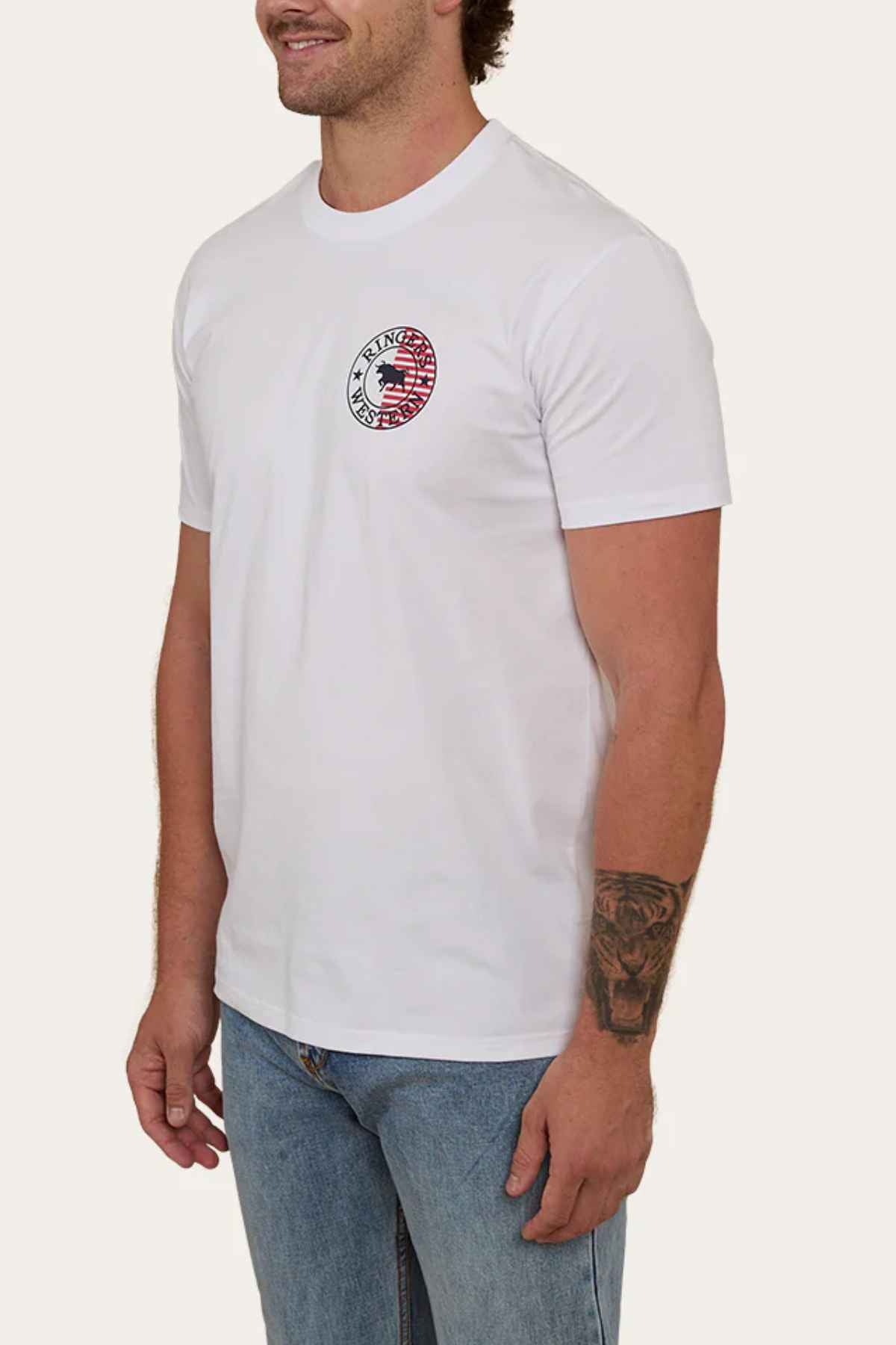 A man wears a Ringers Western Signature Flag Stock Fit T-Shirt in white, made from cotton jersey and featuring a small circular graphic in red, white, and black on the chest. He pairs the T-shirt with light blue jeans and reveals an arm tattoo. His left hand rests at his side as he offers a slight smile.
