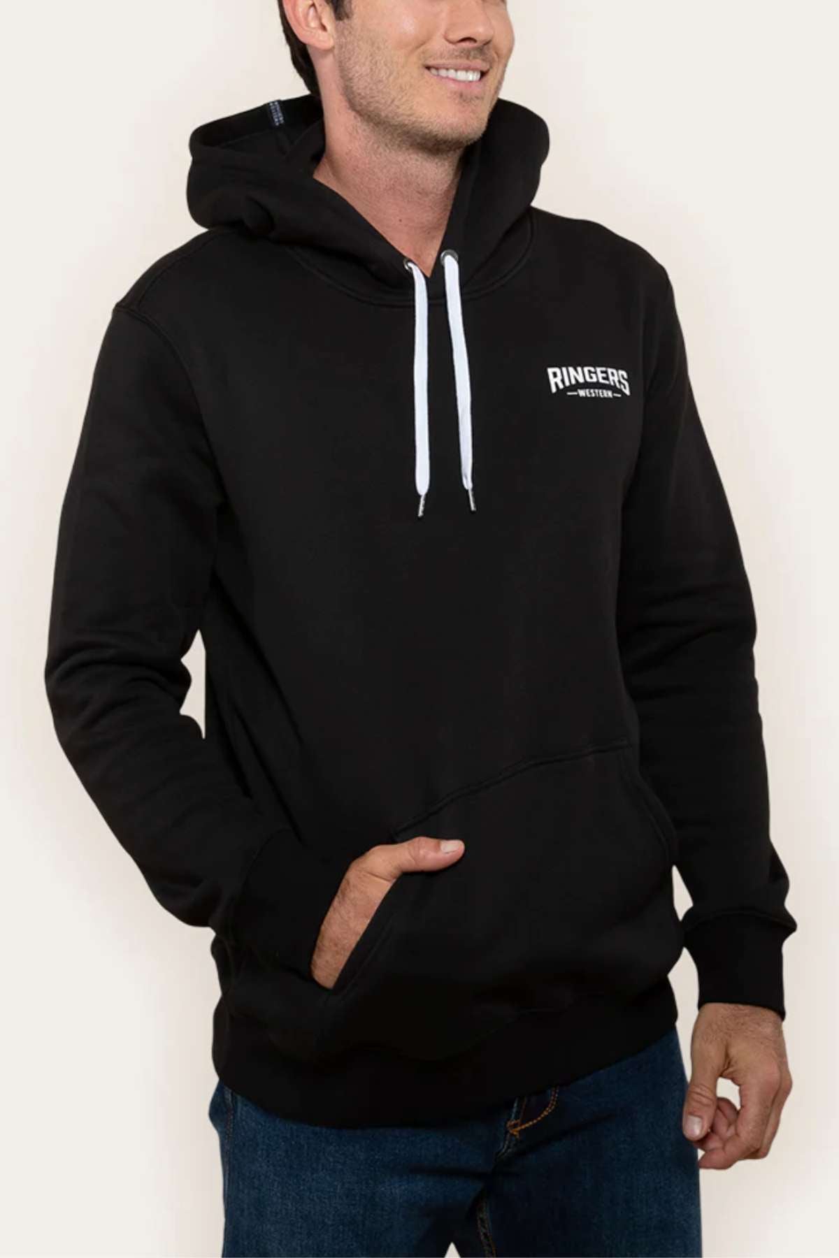 An individual is seen smiling while wearing a black Ringers Western Squadron Hoodie made of cozy brushed fleece. The hoodie has the brand name printed on the left chest, features a kangaroo pocket, and is accented with white drawstrings. The person has their right hand in the pocket while their left hand rests by their side.