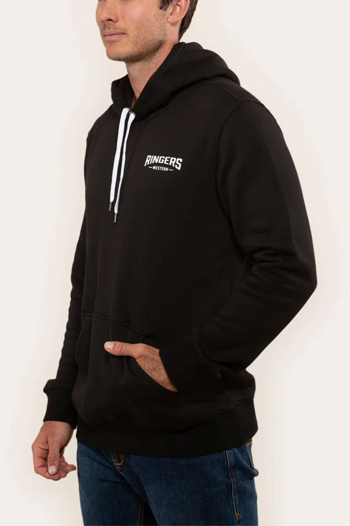 A person wearing a Ringers Western Squadron Hoodie in black, characterized by white drawstrings and a subtle white logo on the chest. The brushed fleece sweatshirt features a convenient kangaroo pocket where they have one hand tucked in, paired with blue jeans. The backdrop is plain and light-colored.