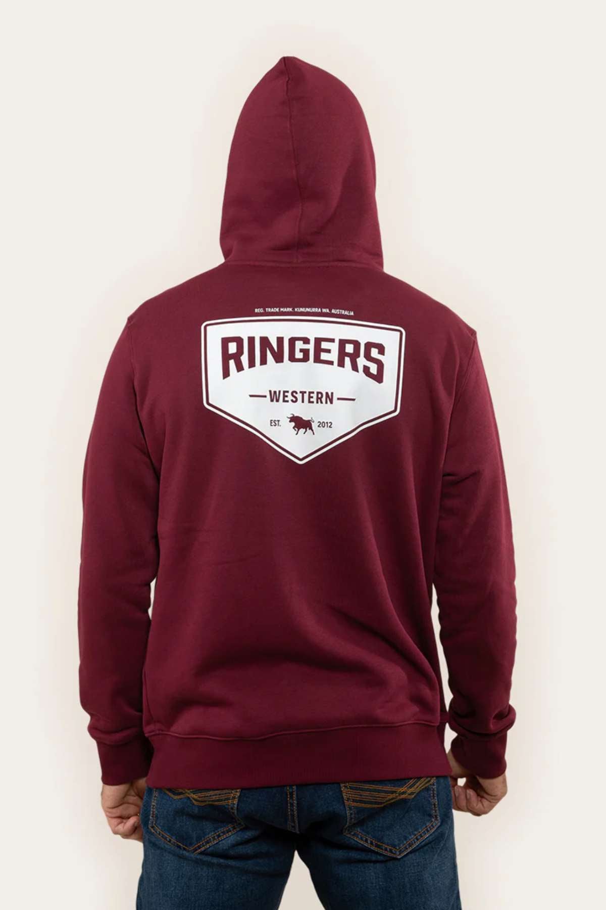 A person wearing the Ringers Western Squadron Hoodie in a relaxed fit and maroon color stands facing away from the camera. The hoodie showcases a large white shield-shaped logo on the back that reads "Ringers Western Est. 2012," along with a small bull graphic at the bottom of the logo. The person is also wearing jeans.
