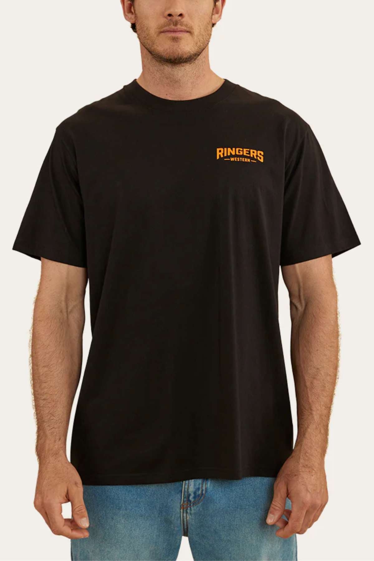A person is wearing a Ringers Western Squadron Loose Fit T-Shirt in plain black, made from 100% cotton jersey with "RINGERS WESTERN" printed in orange on the left side of the chest. The pre-shrunk t-shirt is paired with light blue jeans. The background is a plain, off-white color.