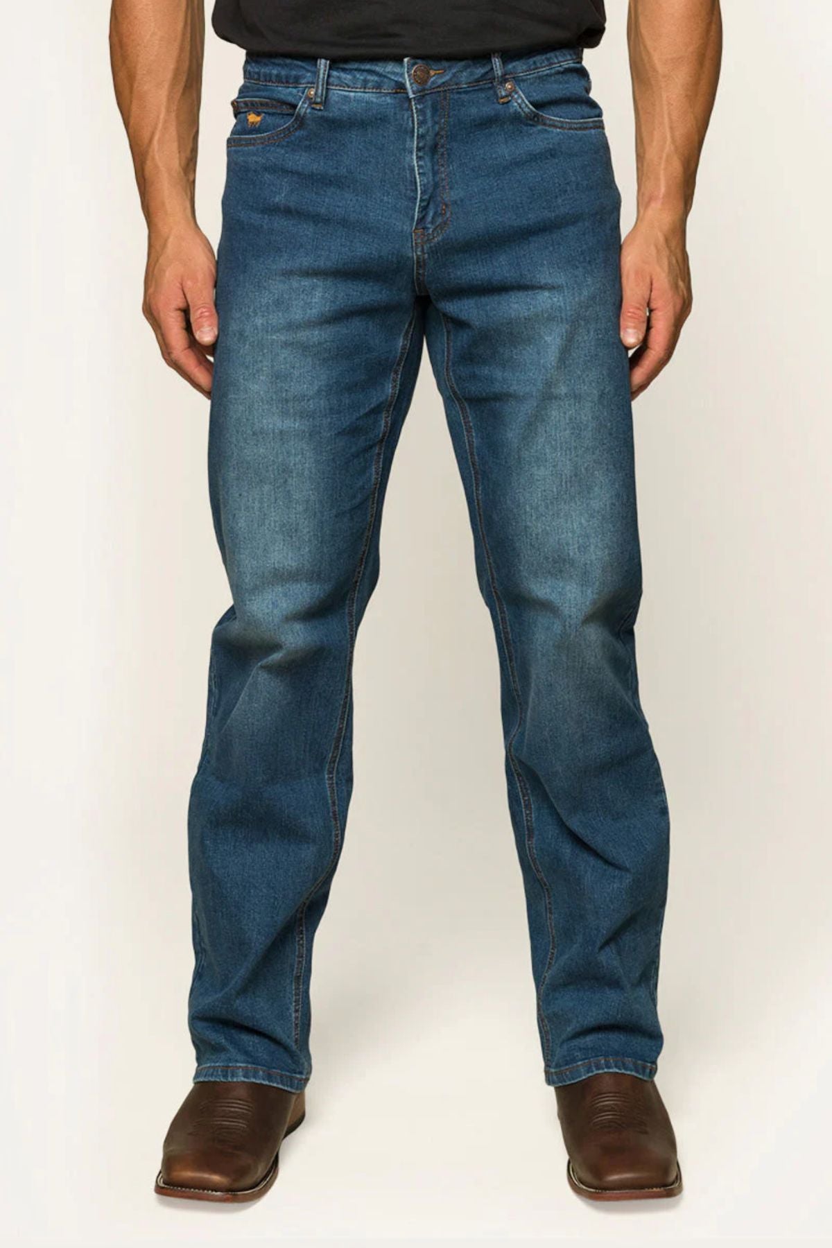 A man in Ringers Western Sturt Mens Relaxed Leg Jeans and brown boots stands against a neutral backdrop. The jeans, crafted from premium stretch denim, have a classic cut with visible stitching and slight fading on the thighs and knees. The lower part of a dark shirt is visible.