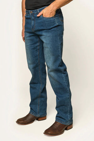 A person in Ringers Western Sturt Men's Relaxed Leg Jeans, with premium stretch denim and slight fading on the thighs and knees, stands with one hand in a pocket. Polished brown cowboy boots with visible stitching complete the look.