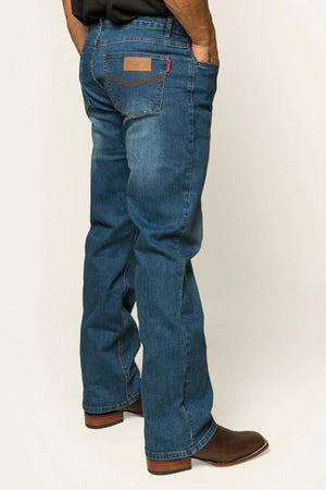 A man in Ringers Western Sturt Mens Relaxed Leg Jean and brown leather boots has his hands in his pockets. The jeans, made from premium stretch denim, feature a visible back pocket with a brown patch against a plain neutral background.