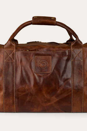 The Ringers Western Toro Gino Overniter is a brown full grain leather duffel with sturdy handles, visible stitching, and a square front patch embossed with the logo—your ideal durable travel companion.