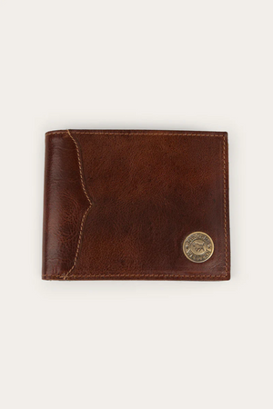 The Ringers Western Toro Grande Wallet is a brown full grain leather wallet with visible stitching and a round metal emblem in the bottom right corner, shown closed on a plain light background.