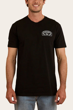 A man in a black 100% cotton Ringers Western Wheatbelt Stock Fit T-Shirt with a small white left-chest logo, paired with light blue jeans, stands against a plain light background.