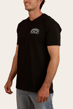 A man in the Ringers Western Wheatbelt Stock Fit T-Shirt, crafted from 100% cotton, pairs it with light blue jeans and stands smiling against a plain background.