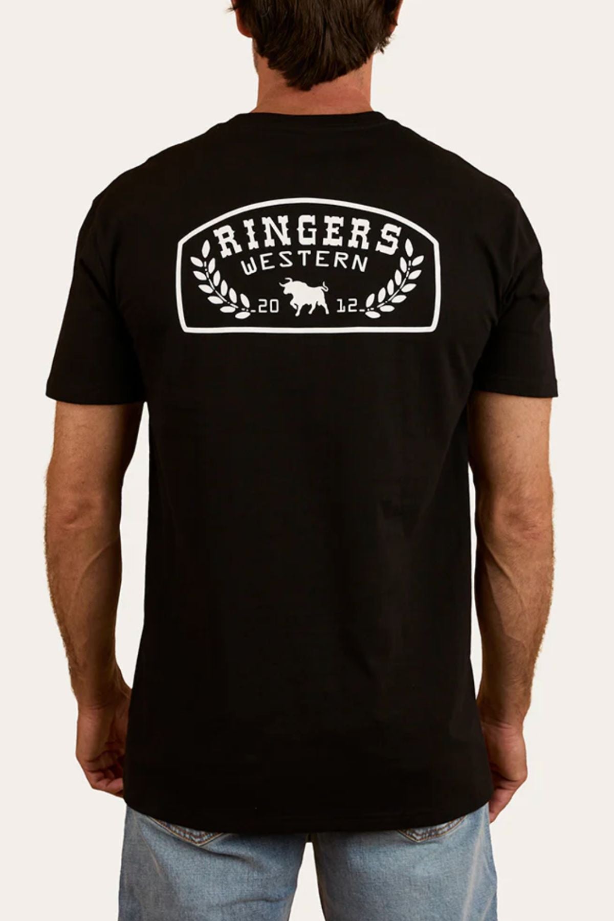 Someone stands facing away, hands at their sides, wearing a black Ringers Western Wheatbelt Stock Fit T-Shirt made from 100% cotton, featuring a bold white bull logo and "Ringers Western" on the back.