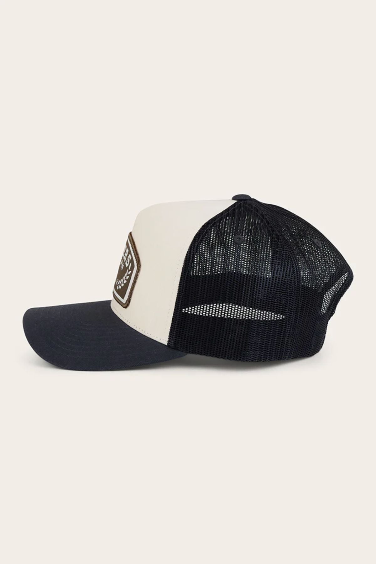 The Ringers Western Wheatbelt Trucker Cap features a curved bill, white cotton twill front, black mesh back, and a brown patch on the front. Shown from the side against a light background.