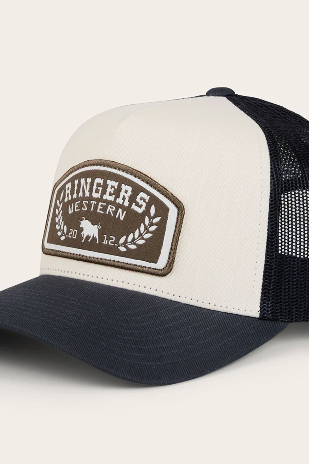 The Ringers Western Wheatbelt Trucker Cap features a cream and black design with a mesh back, cotton twill front, and a patch displaying "Ringers Western," a laurel wreath, bull graphic, and "2012" on each side of the wreath.