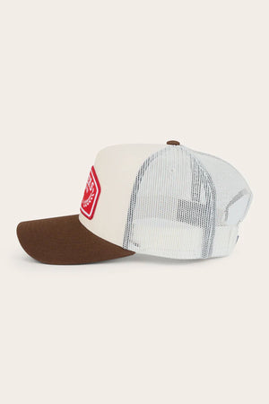 The Ringers Western Wheatbelt Trucker Cap features a brown bill, white front panel with a red patch, and breathable mesh back. Made from cotton twill, it's shown side-on against an off-white background.