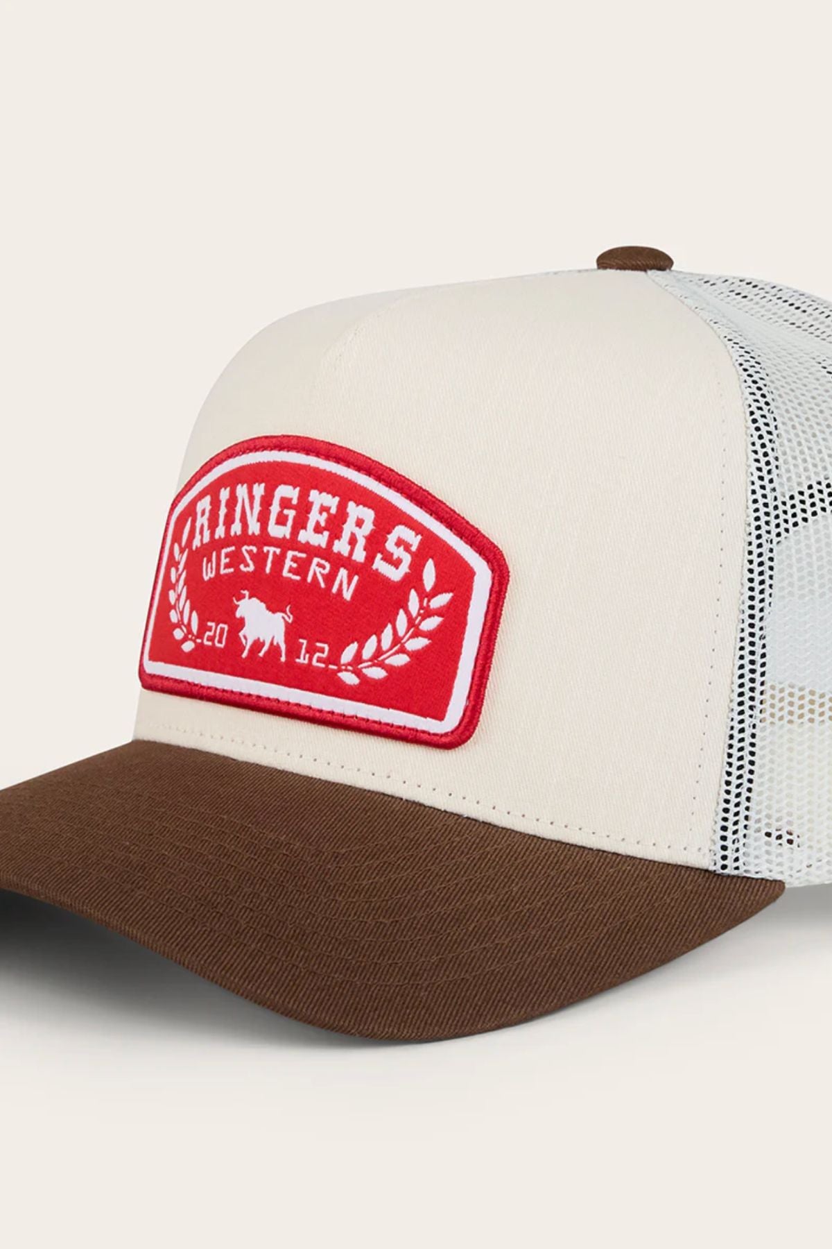 The Ringers Western Wheatbelt Trucker Cap features a beige and brown design, white breathable mesh back, and a red patch with "RINGERS WESTERN 2012," a small bull, and laurel wreath.