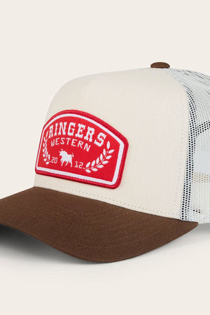 The Ringers Western Wheatbelt Trucker Cap features a beige and brown design, white breathable mesh back, and a red patch with "RINGERS WESTERN 2012," a small bull, and laurel wreath.
