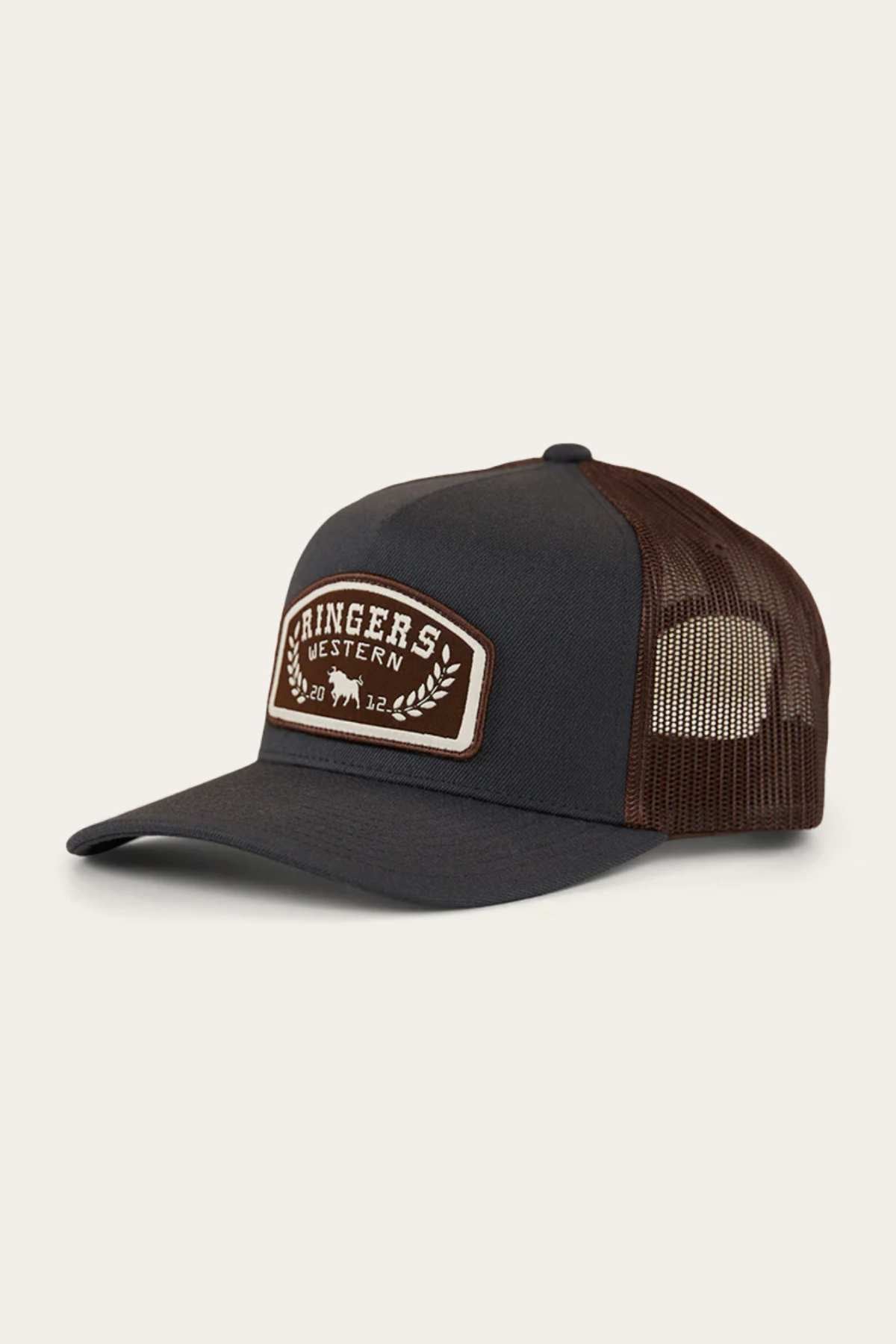 The Ringers Western Wheatbelt Wool Trucker Cap features a dark grey and brown design with a curved brim and adjustable strap, adorned with a patch showing decorative leaves and two cattle silhouettes. It has breathable brown mesh fabric on the sides and back.