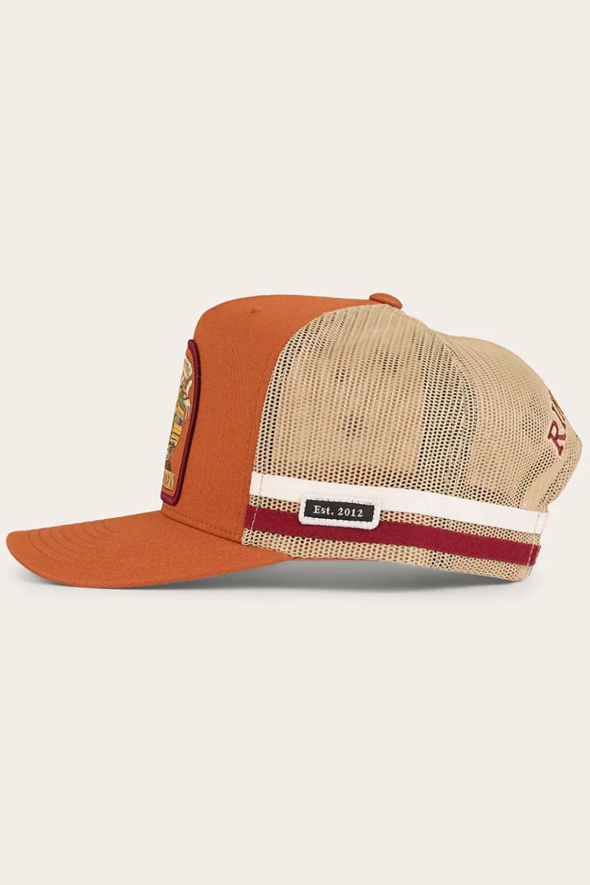 A side view of the Ringers Western Workhorse Trucker Cap reveals its burnt orange front, tan mesh back for breathability, a front patch, striped back band, and a label reading "Est. 2012".