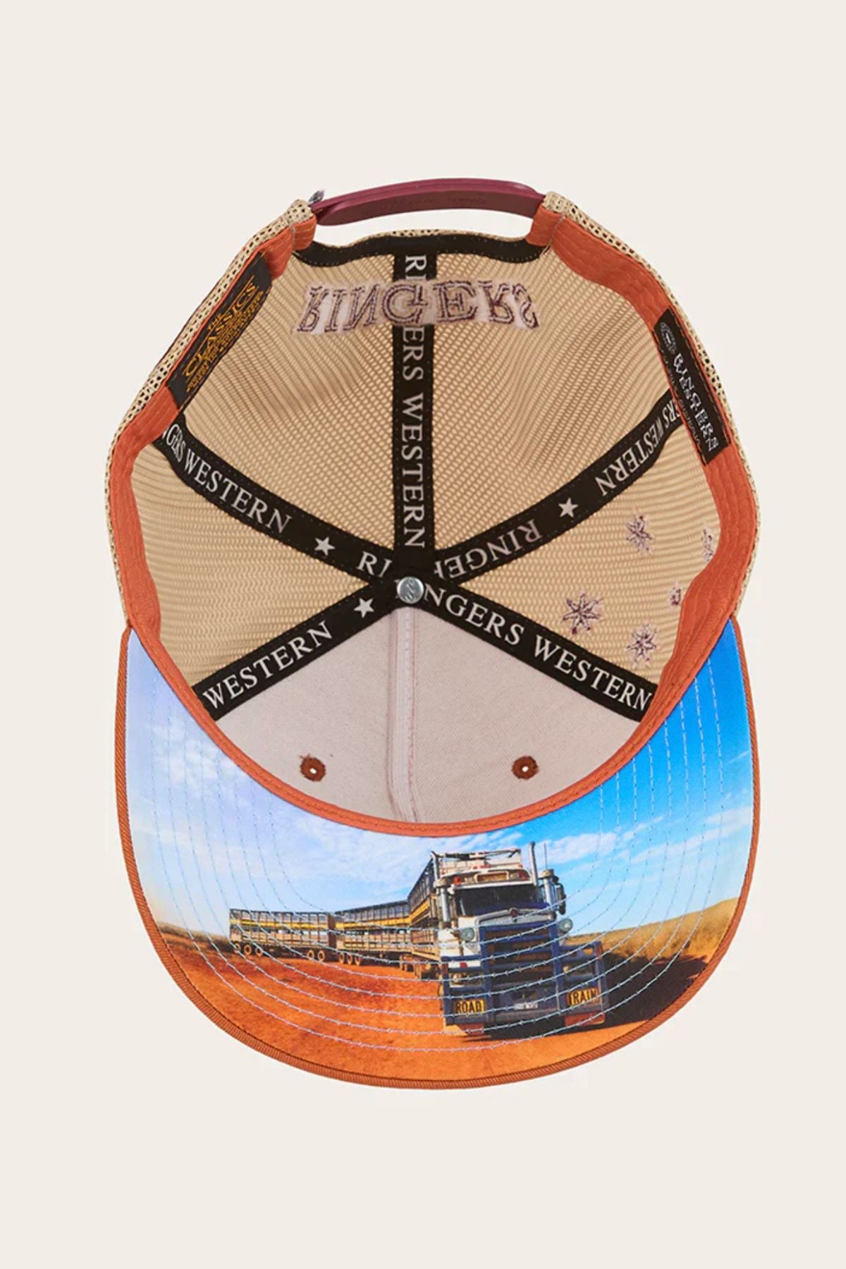 The Ringers Western Workhorse Trucker Cap is shown upside down, revealing the inner lining with "RINGERS WESTERN" text. Its brim features a truck on a dirt road in a rural landscape, combining classic trucker style and sun protection.