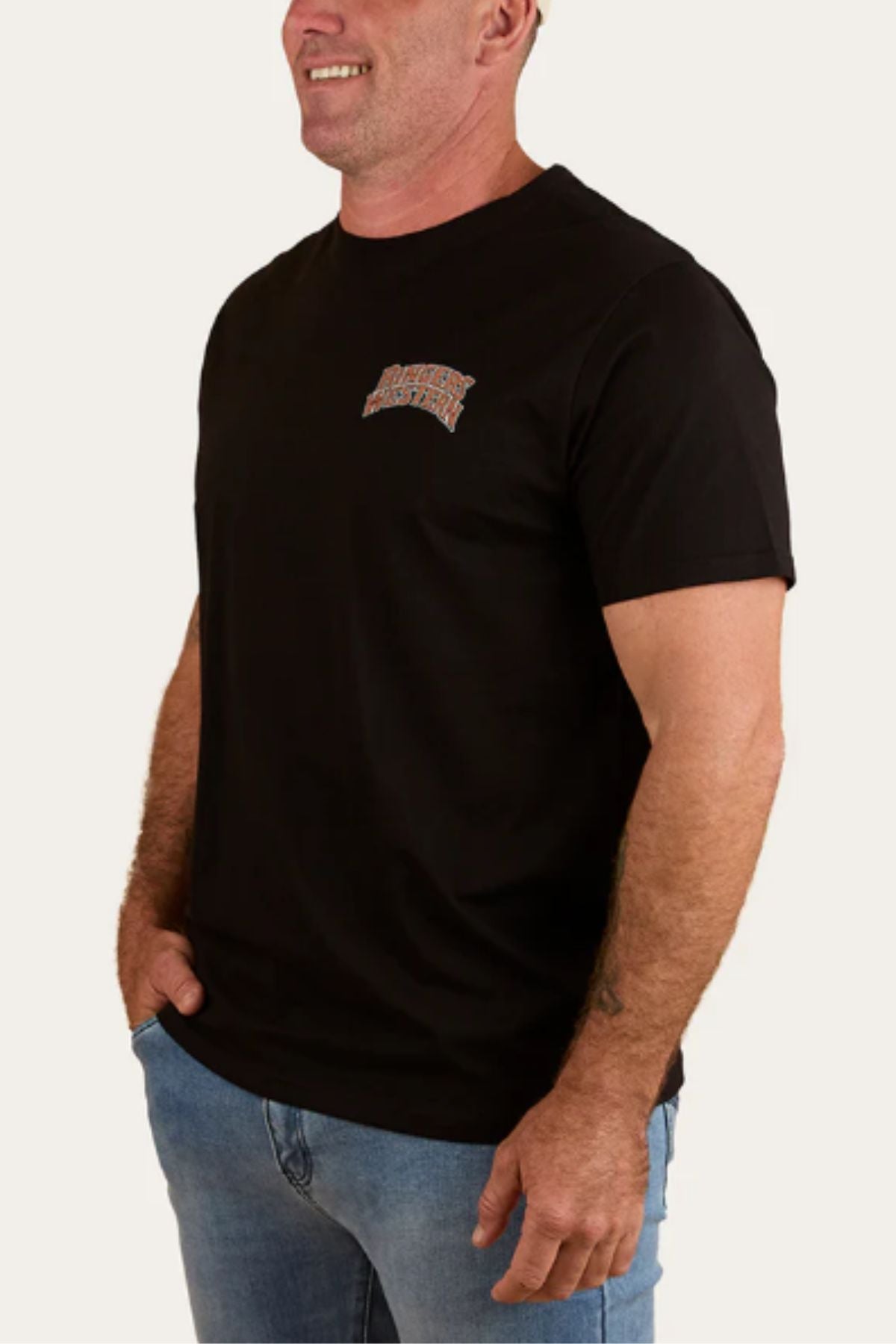 A man smiles with his left hand in his pocket, wearing the Ringers Western Do No Harm Take No Bull Loose Fit T-Shirt—a black short-sleeve jersey with a colorful left chest graphic—paired with light blue jeans against a plain background.